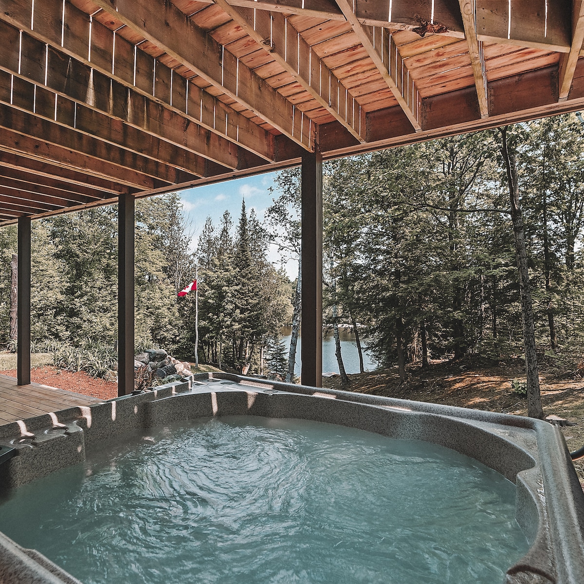 A hot tub is positioned on a covered outdoor deck, surrounded by trees and offering a view of the lake. Natural light filters through the structure, creating a serene atmosphere while the Canadian flag gently flutters in the background.