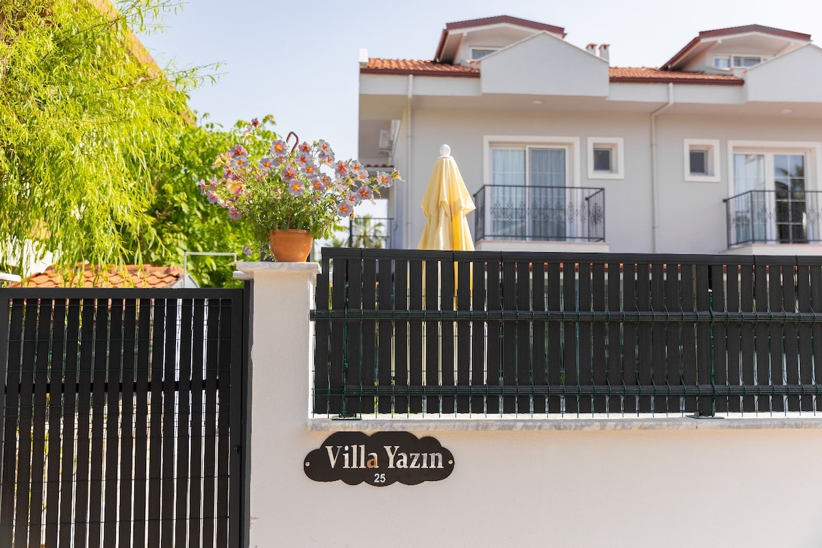 A charming entrance features a black wooden fence with a decorative sign reading 'Villa Yazın' affixed to it. Potted flowers add a touch of color, and a yellow umbrella is visible above, providing a hint of shade to the area. The villa's two-story structure is seen in the background.