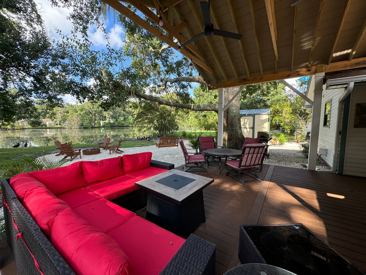 A covered deck features a seating area with bright red cushions surrounding a gas fire pit. The serene pond is visible nearby, framed by lush greenery. In the background, additional seating arrangements are discernible along pebbled pathways.