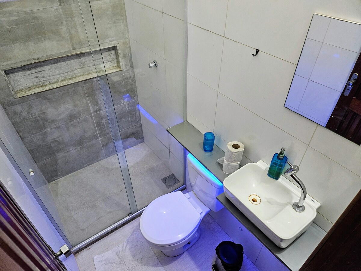 A modern bathroom is equipped with a glass shower, toilet, and sink. Light blue accents are present under the countertop. A mirror is mounted on the wall, and toiletries are neatly arranged on the counter, contributing to a functional and clean atmosphere.