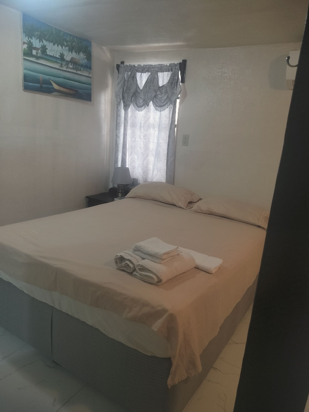 A well-lit sleeping area features a spacious bed with a neutral-colored bedspread. Soft towels are neatly arranged at the foot of the bed. A window, dressed with semi-sheer curtains, allows natural light to fill the space, while a painting is displayed on the wall.