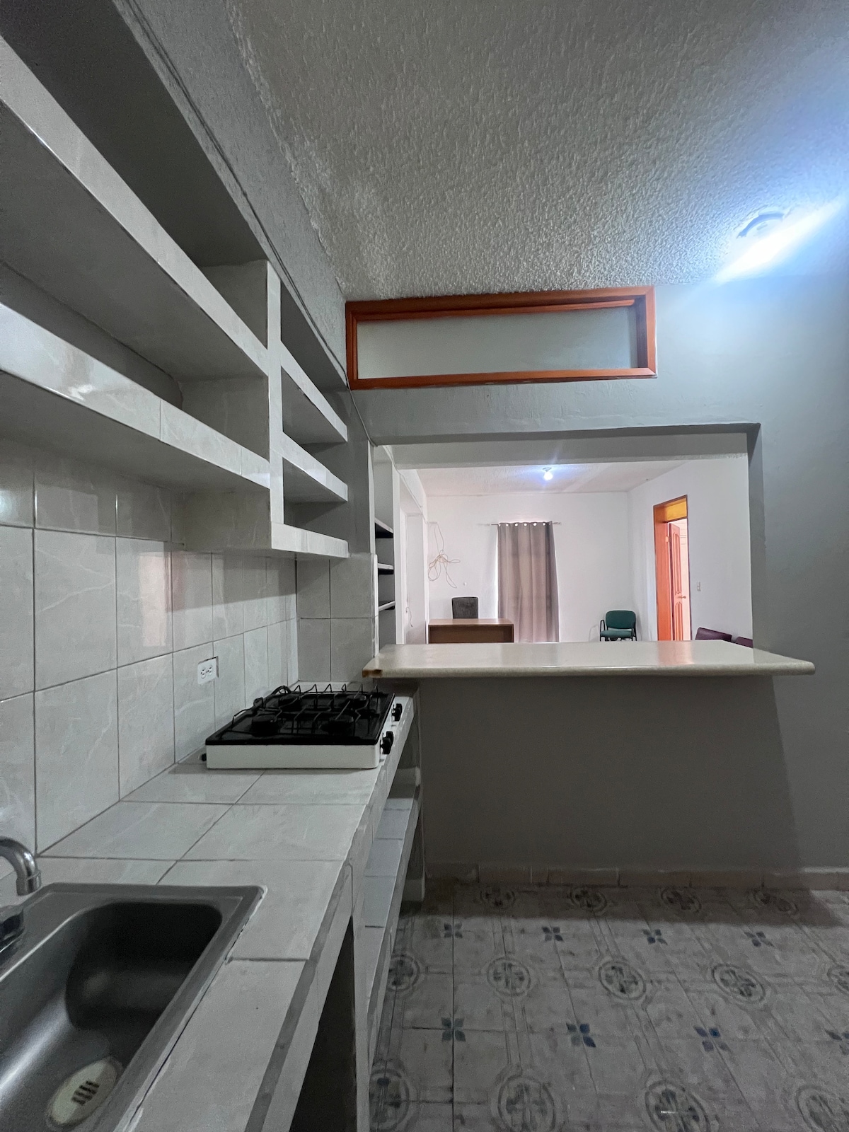 A well-organized kitchen features light-colored cabinetry and a spacious countertop. A gas stove is positioned against the wall, with a kitchen sink located beneath an open shelf. Natural light is present, illuminating the adjacent area where a living space can be seen.
