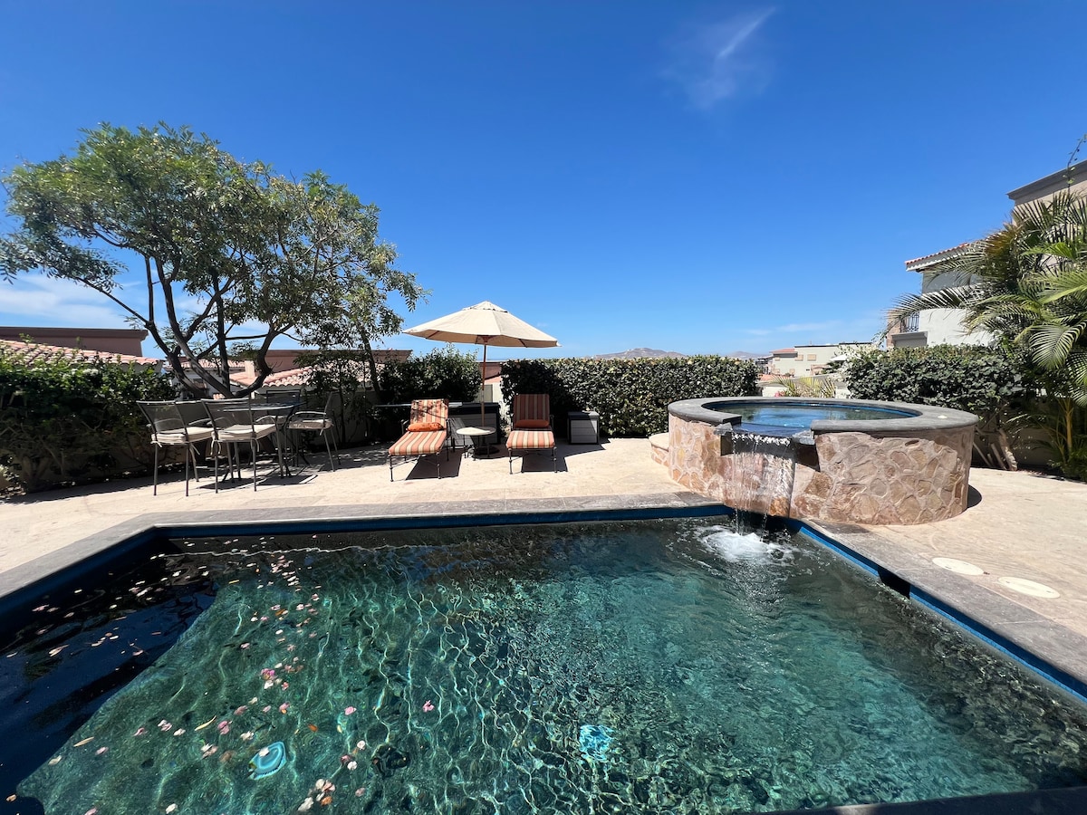 An outdoor area features a sparkling pool with a built-in spa, surrounded by a stone patio. Comfortable lounge chairs are positioned under an umbrella, accompanied by a dining set. Lush greenery adds a natural touch, creating a relaxing atmosphere.