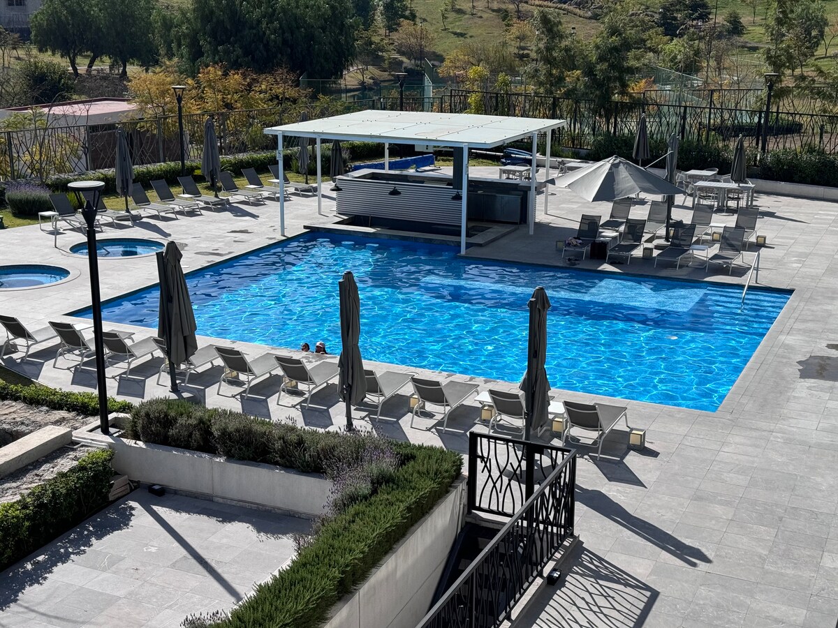 A large swimming pool is surrounded by lounge chairs and umbrellas, providing ample space for relaxation. A shaded cabana area is visible, offering seating near the pool. The area is well-maintained, with greenery lining the edges and a backdrop of gentle hills.