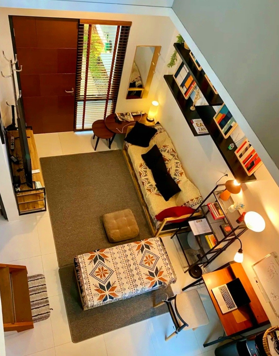 An overhead view captures a well-arranged living space with a cozy sofa adorned with decorative pillows. A small table sits nearby, accompanied by a compact workspace featuring a laptop. Shelves are lined with books, while a warm light provides illumination within the serene atmosphere.