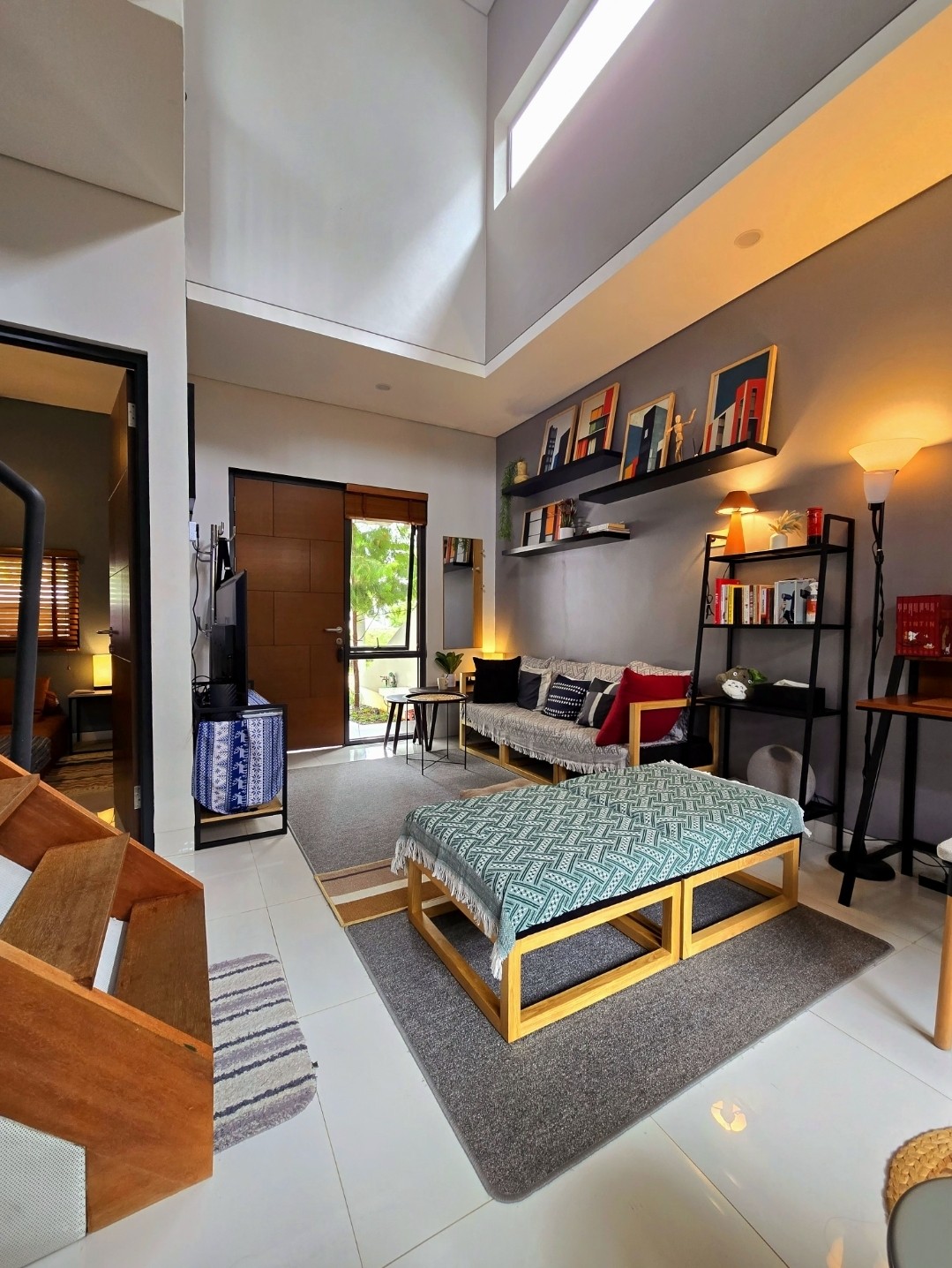 A cozy living area features a comfortable seating arrangement with a patterned throw on a low platform. A tall bookshelf displays various books, while an entry door allows natural light to enter. Soft lighting creates a welcoming ambiance, complemented by a small desk in the corner.