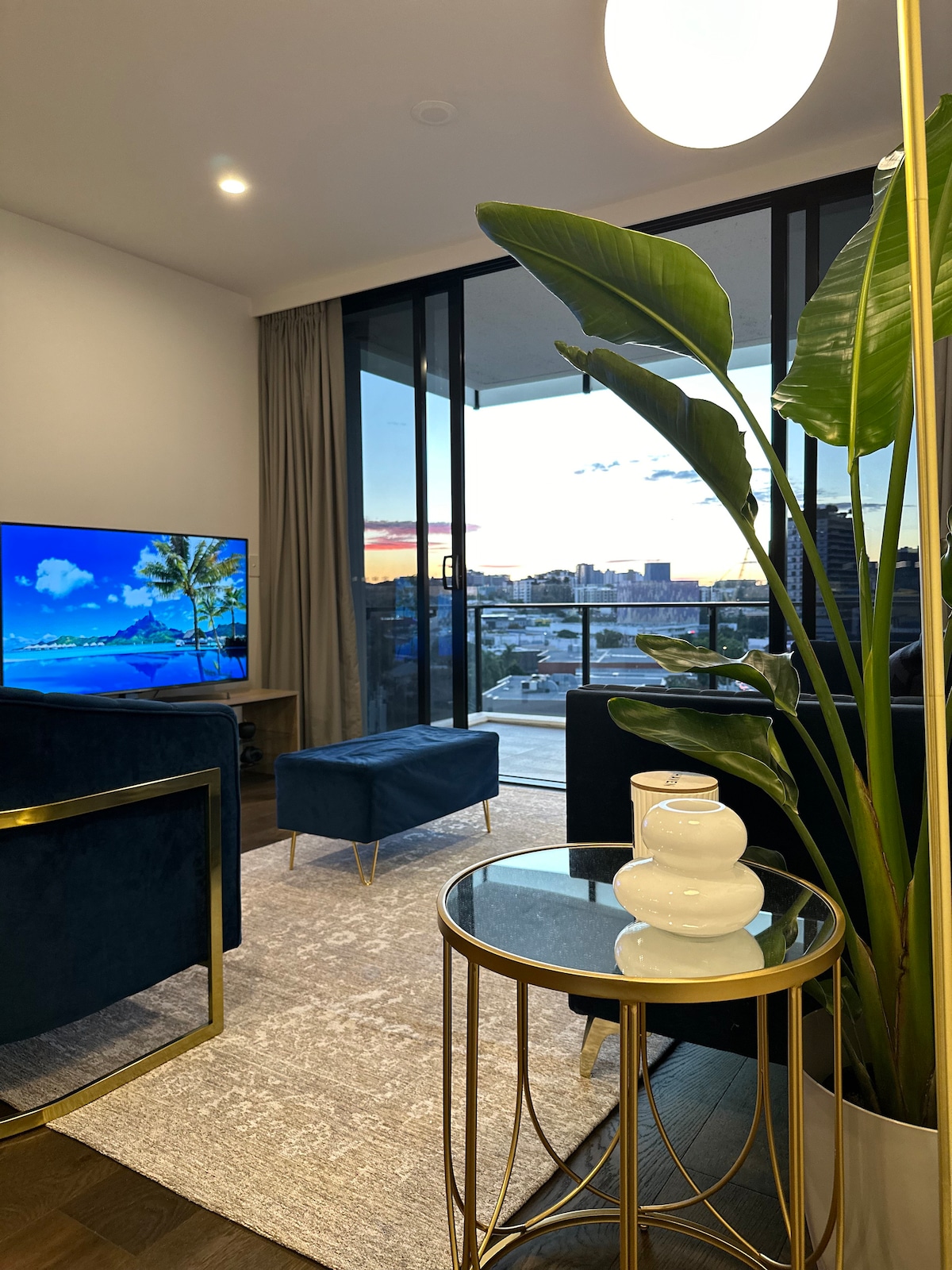 A modern living area is shown with two plush chairs and a small side table. A large window offers city views, while a green plant adds a touch of nature. A geometric rug lies on the floor, complementing the warm lighting from a glass pendant lamp.