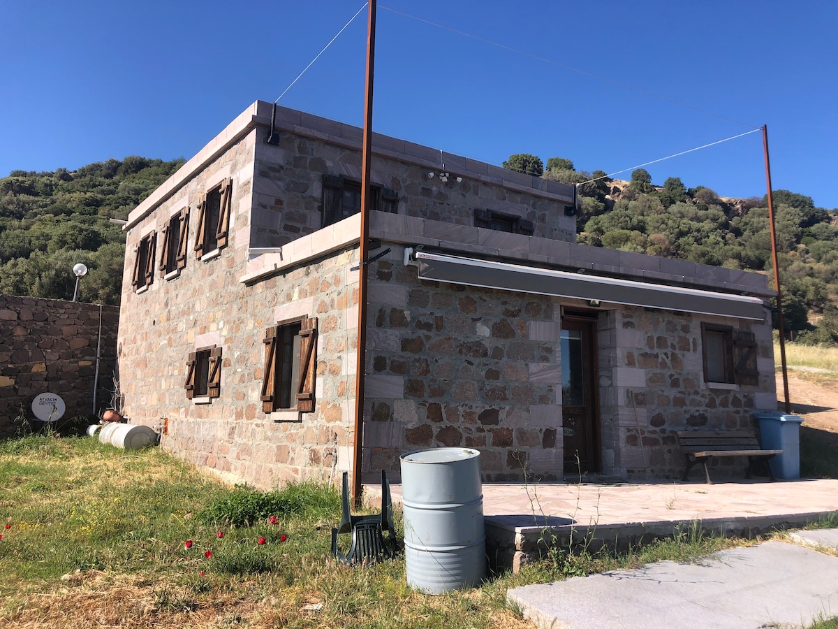A two-story stone building is displayed, featuring wooden shutters and a simple porch area. The surrounding landscape includes green grass and distant hills. A pathway leads to the entrance, while barrels are positioned nearby, contributing to the rustic environment.