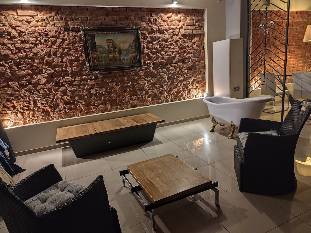 A stylish living area features a rich brick wall illuminated by soft lighting. Two wicker armchairs are positioned beside a wooden coffee table, providing a comfortable seating arrangement. A vintage bathtub adds a unique touch to the space, enhancing the overall aesthetic.
