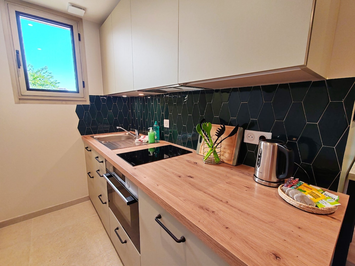 A modern kitchen space features sleek cabinetry and a wooden countertop. A kettle and kitchen accessories are neatly arranged. The hexagonal tile backsplash in deep green adds visual interest. Natural light enters through a window, illuminating the clean and fresh environment.