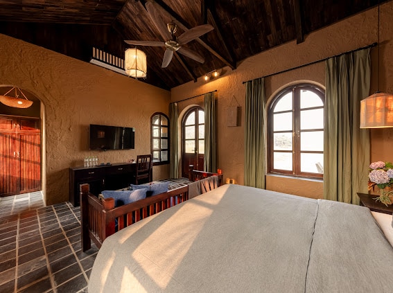 A spacious room features vaulted wooden ceilings and large arched windows allowing natural light to enter. A comfortable bed is complemented by dark wooden furniture, including a seating area and a television. Light, flowing curtains frame the windows, adding a soft touch to the decor.