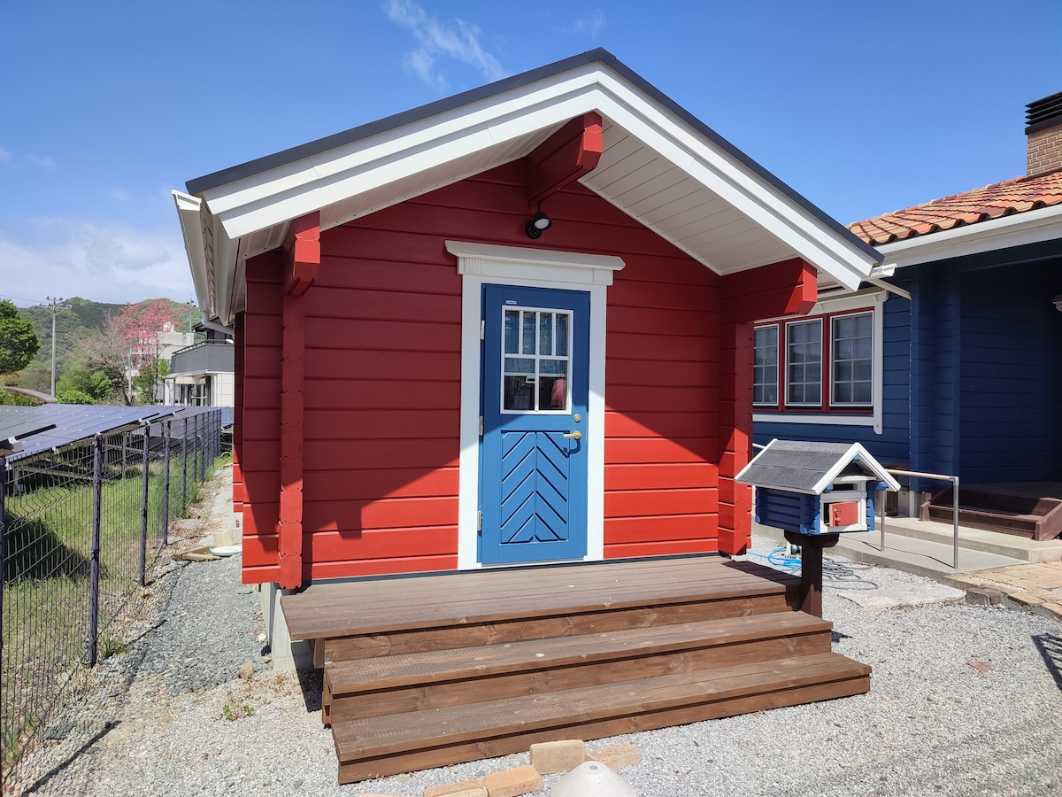 A red wooden cabin with a blue door is showcased, featuring a small mailbox beside the entrance. The front porch is made of wooden decking, and the surrounding landscape includes a fence and greenery in the background.