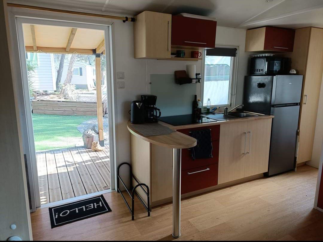 A compact kitchen area is equipped with modern cabinetry and appliances, including a coffee maker and refrigerator. A curved breakfast bar offers seating, while a door leads to a wooden deck area outside, framed by greenery.