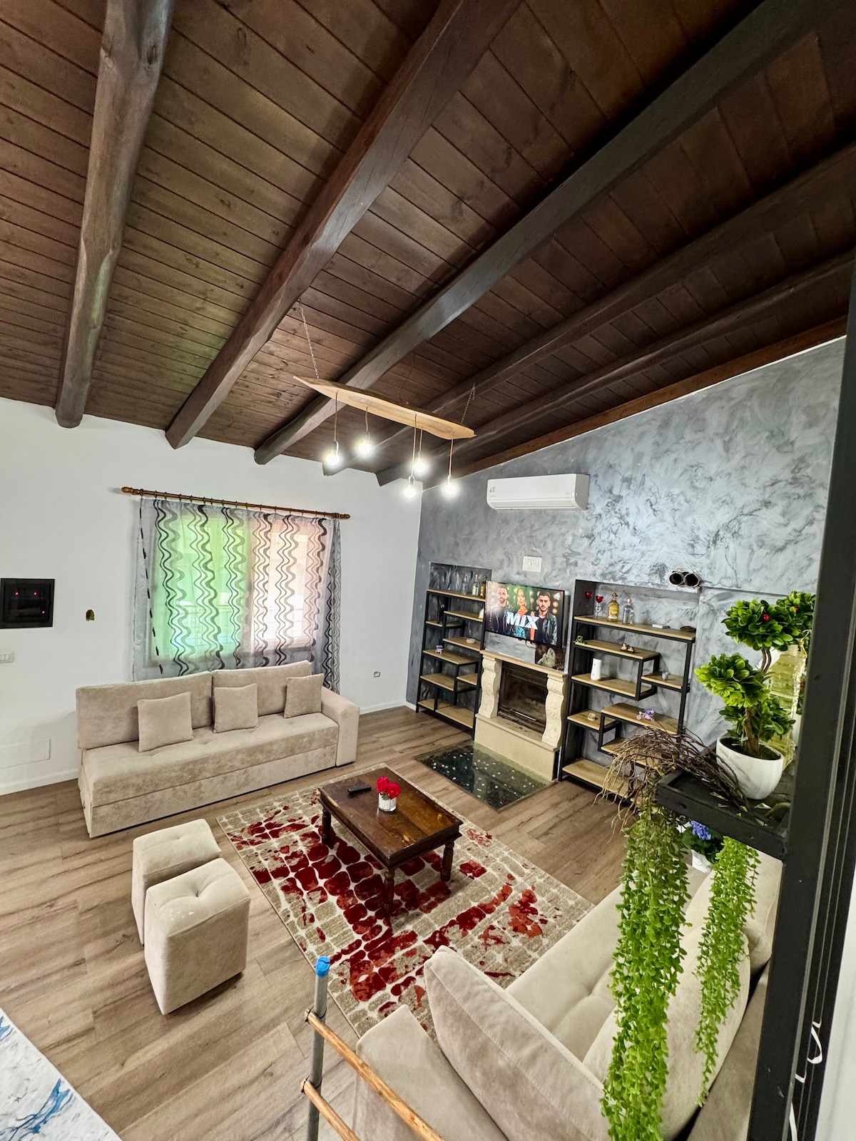 A spacious living room is presented, featuring a soft seating arrangement with light-colored couches and ottomans. A wooden coffee table occupies the center, while a modern television and decorative shelving are visible against one wall. Natural light filters through patterned curtains, enhancing the inviting ambiance.