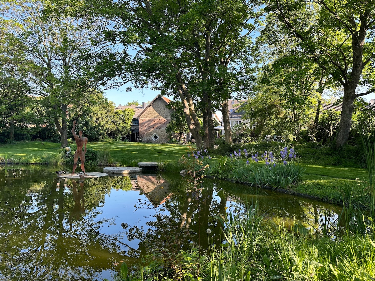 A serene garden is depicted, featuring a tranquil pond that reflects the surrounding trees and house. The area is adorned with greenery and colorful flowers, enhancing the peaceful setting. A sculpture stands in the foreground, adding an artistic element to the natural landscape.