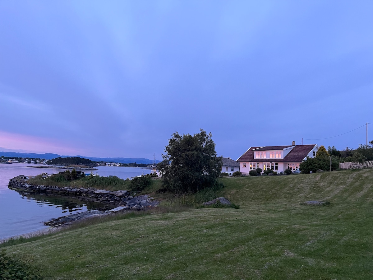 The exterior of a newly renovated house is captured during twilight, with soft colors reflecting on the windows. A spacious lawn with manicured grass leads to the water's edge, framed by gentle hills and a serene coastline. The peaceful atmosphere is enhanced by the natural landscape.