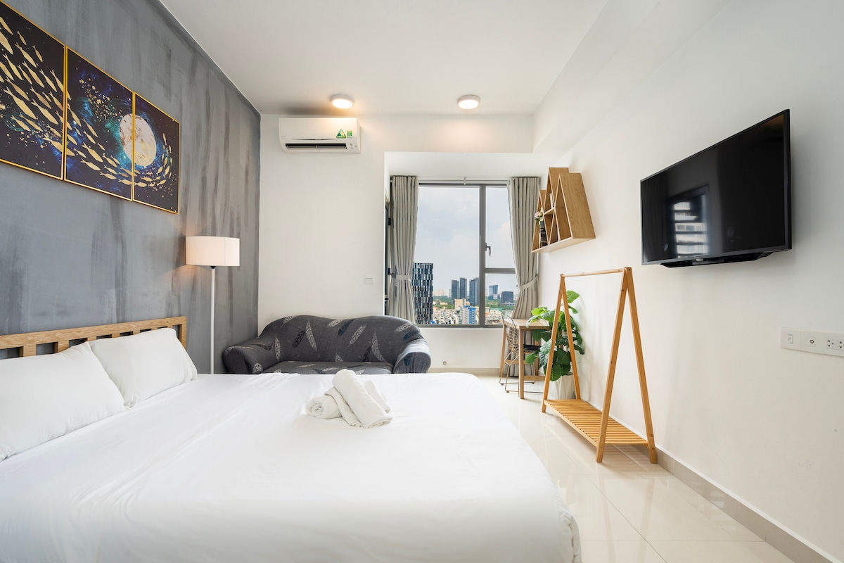 A spacious studio layout features a large bed with clean bedding and soft lighting. A cozy sofa is positioned against the wall, adjacent to a wall-mounted flat-screen TV. Large windows provide city views, while decorative wall art adds character to the space.