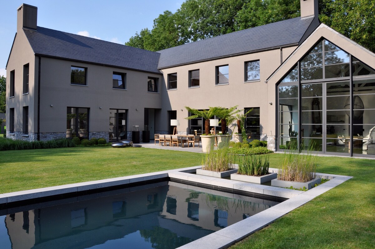 A modern house is depicted with a blend of large windows and a minimalist facade. A landscaped garden surrounds a rectangular swimming pool, reflecting the structure’s design. An outdoor dining area with a wooden table and chairs is visible, enhancing connection with the natural setting.