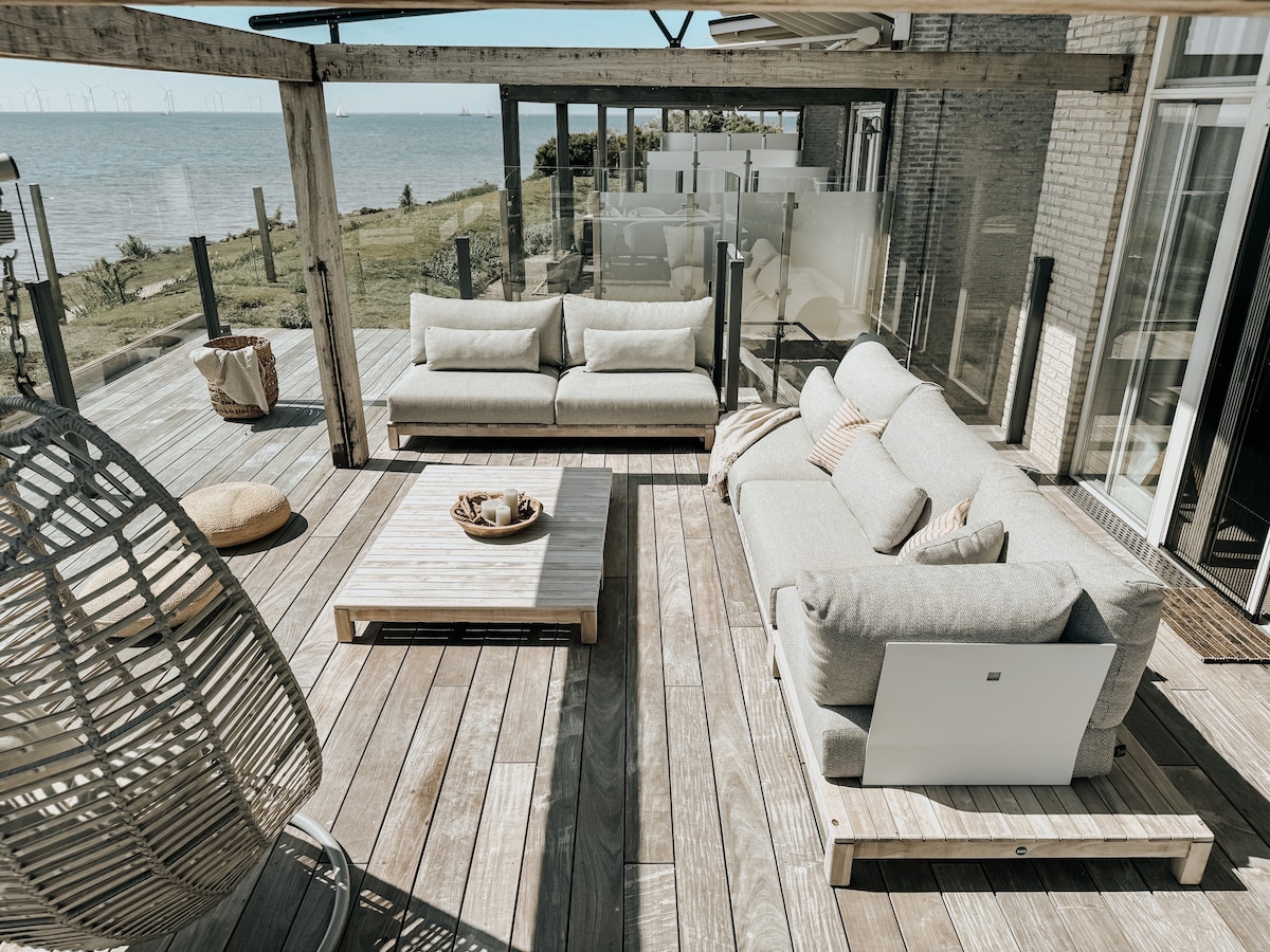 A spacious terrace is furnished with light-colored sofas arranged around a low wooden table. Clear views of the water are offered through the glass panels. A hanging chair adds a cozy touch, surrounded by natural wooden decking, creating an inviting outdoor space.