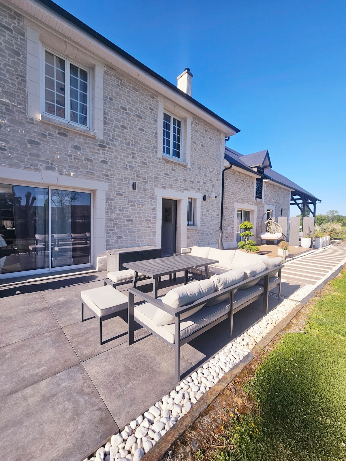 A modern outdoor seating area is depicted, featuring a comfortable sectional sofa accompanied by a matching coffee table. The stone façade of the house is visible in the background, with large windows allowing natural light to enter. The patio area is bordered by green grass and decorative stones.