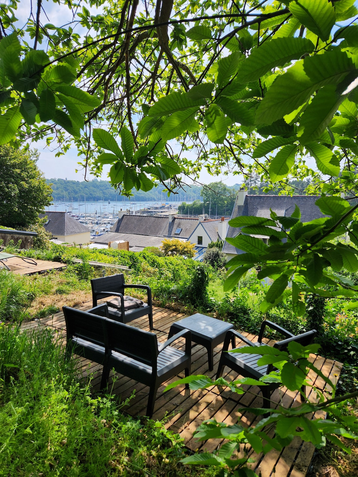 A shaded outdoor seating area is surrounded by lush greenery, featuring a small table and two chairs on a wooden deck. Views of a marina with boats and nearby buildings are partially visible through the leaves of the trees.