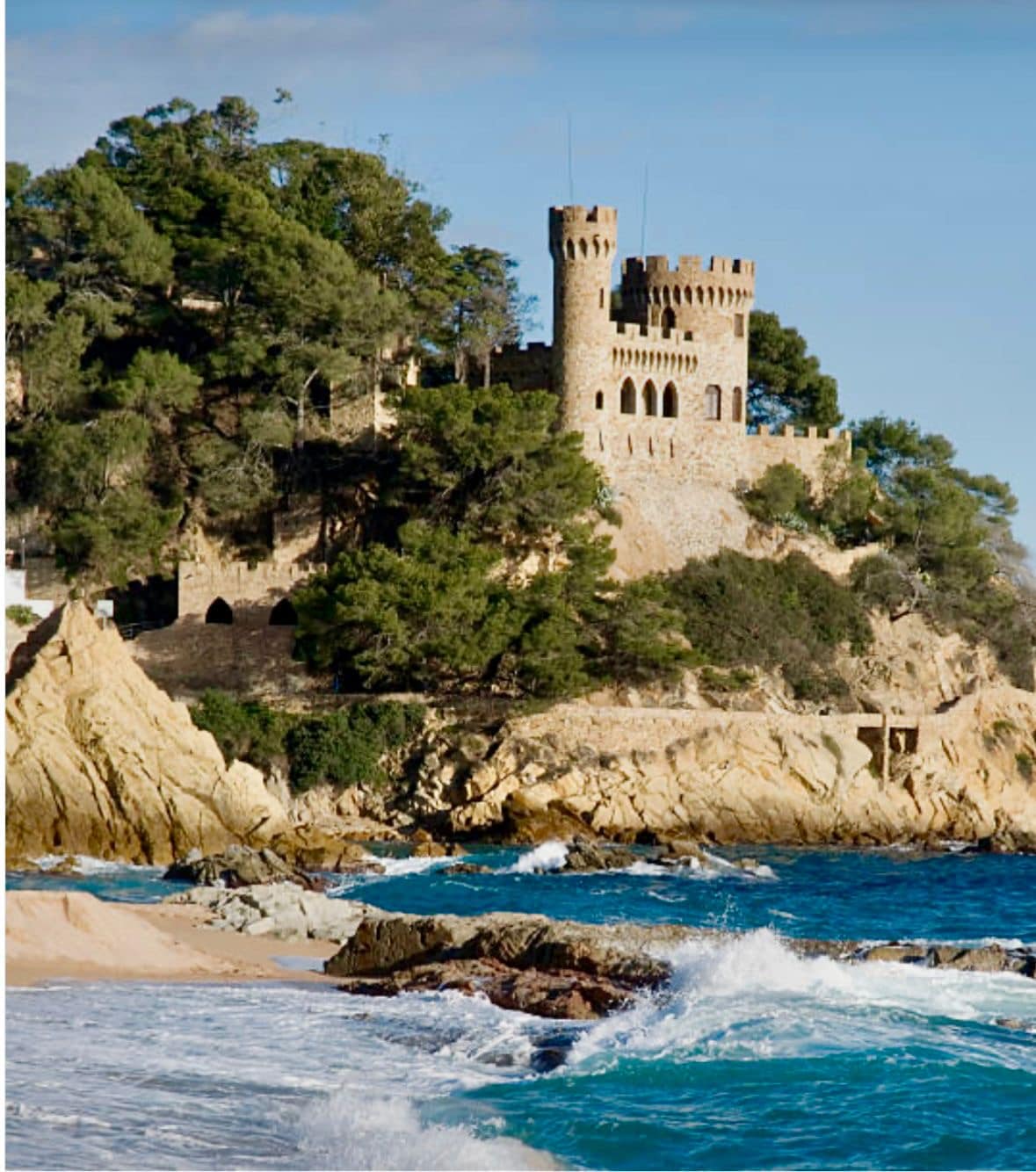 A stone castle is perched atop a rocky cliff, surrounded by lush greenery. The coastline features sandy beaches and waves gently crashing against the rocks below, creating a serene natural landscape under a clear blue sky.