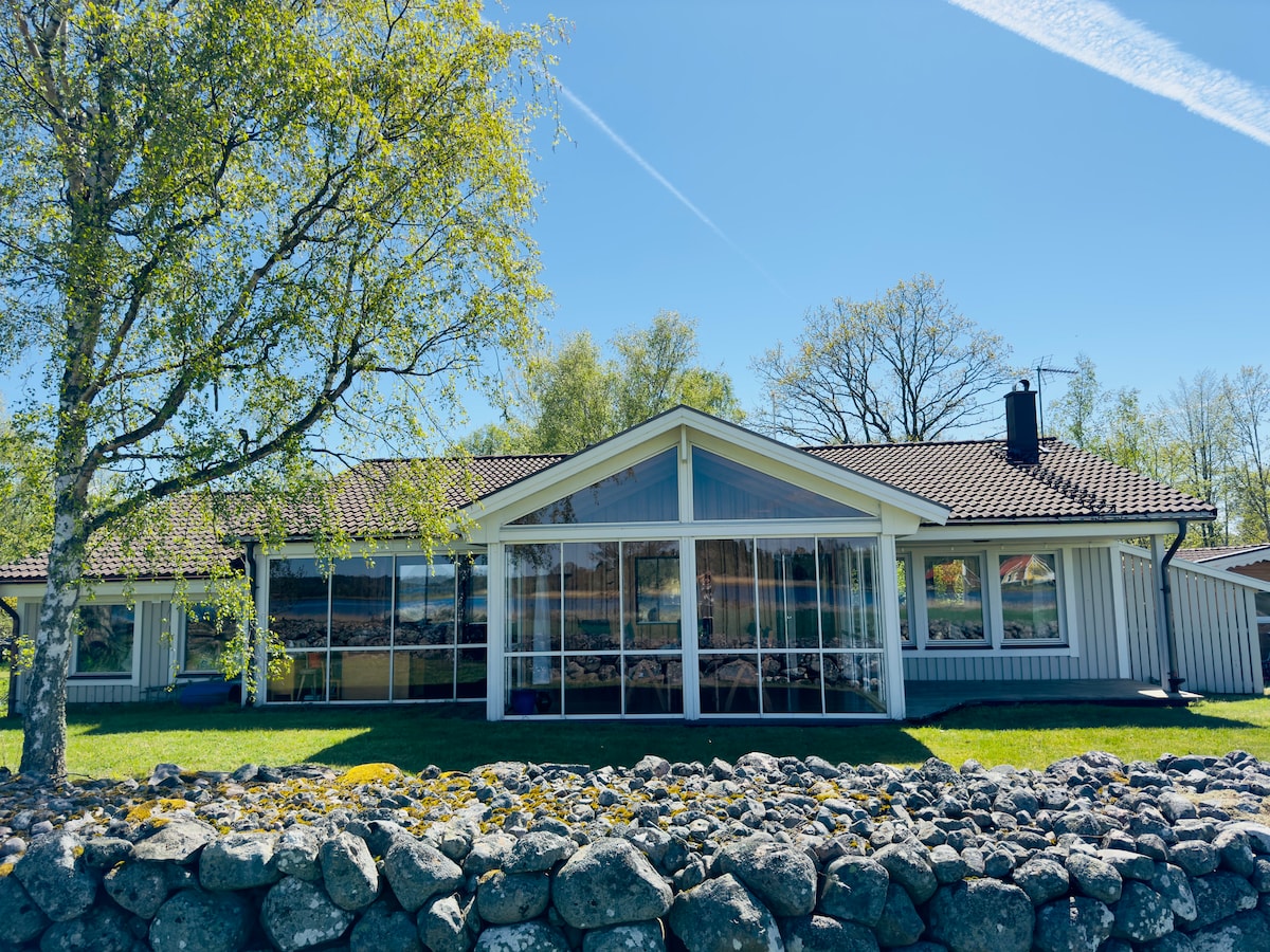 A single-story house features a large front window with multiple panes, allowing natural light to illuminate the interior. A well-maintained lawn surrounds the property, complemented by a stone wall at the foreground. Trees are visible in the background, enhancing the peaceful setting.
