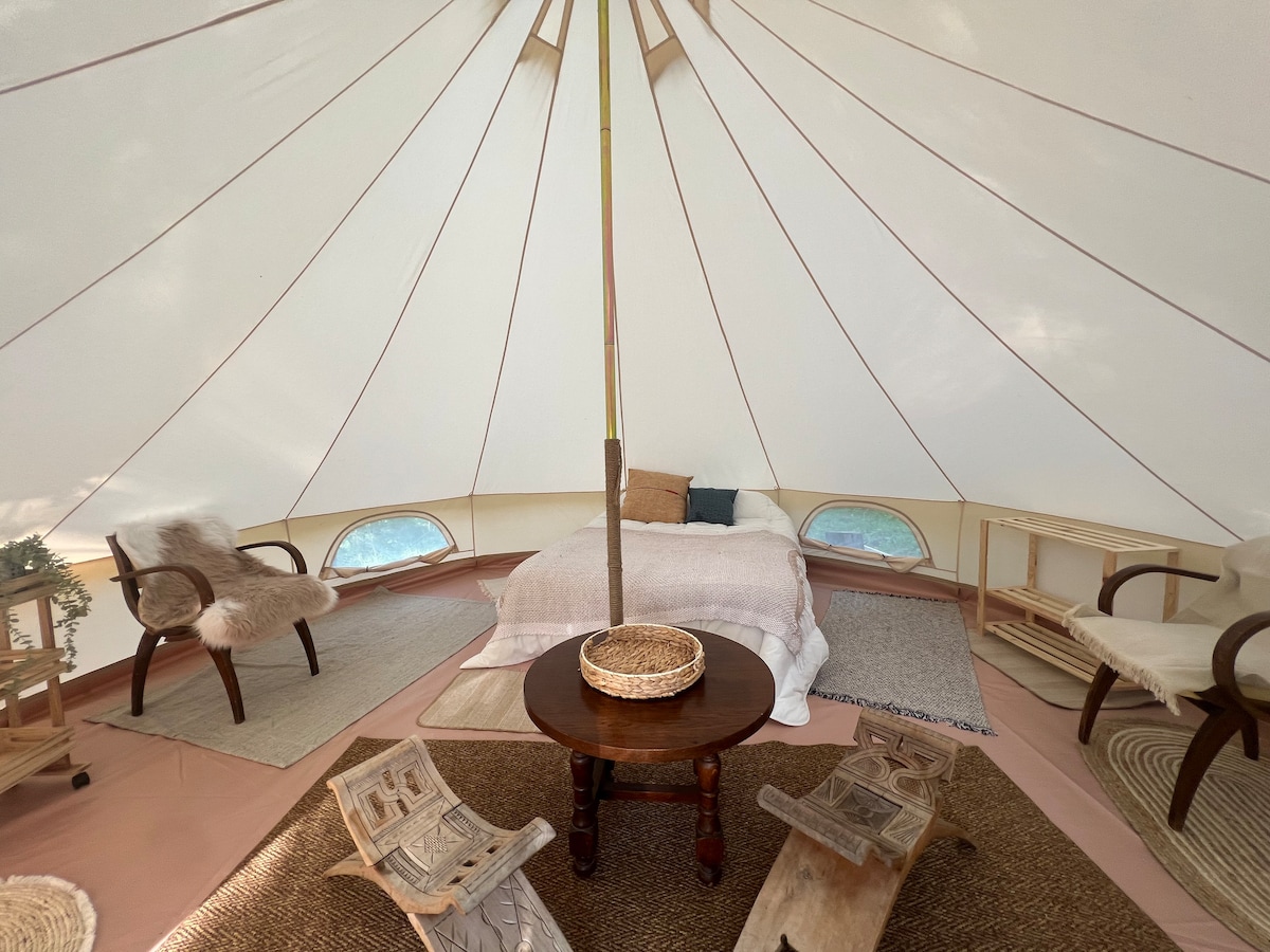 An interior of a spacious tent is captured, featuring a large bed with soft bedding at the center. Surrounding this, two wooden chairs and a round table are positioned on woven rugs. Natural light streams through the tent's windows, highlighting the earthy tones.