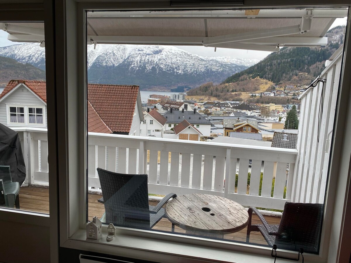 A panoramic view of snow-capped mountains and a serene body of water is framed by large windows. A small round table with two chairs sits on the balcony, offering a cozy spot to enjoy the scenery. Nearby roofs and greenery enhance the tranquil setting.