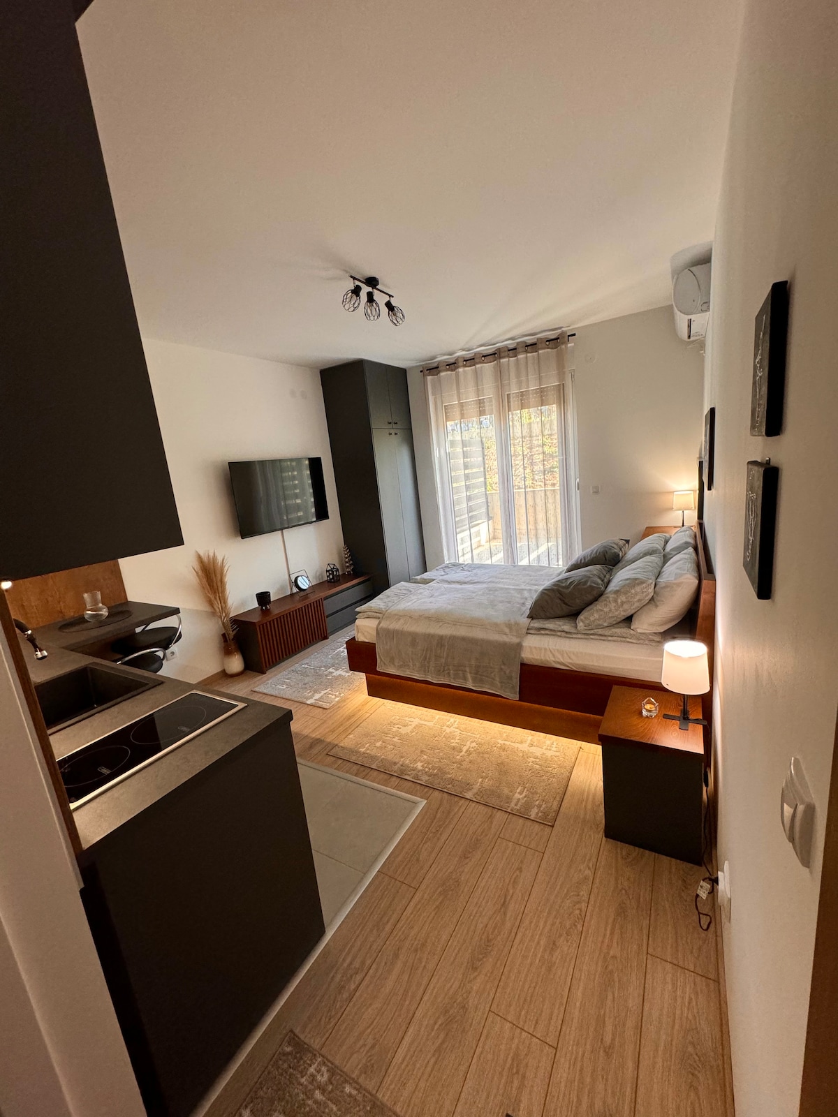 A cozy bedroom area is featured, showcasing a spacious bed with neatly arranged pillows and blankets. A small kitchen area is visible in the foreground, equipped with modern appliances. Natural light filters through sheer curtains, illuminating the warm tones of the furnishings.