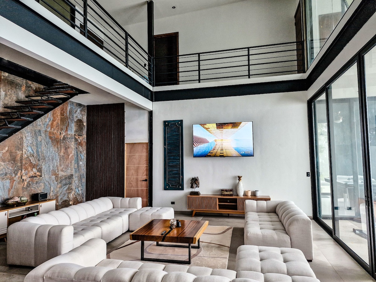 The living area is spacious, featuring high ceilings and large windows that allow natural light to fill the space. A large, light-colored sectional sofa surrounds a wooden coffee table. A flat-screen TV is mounted on the wall opposite the seating, complementing the modern aesthetic.