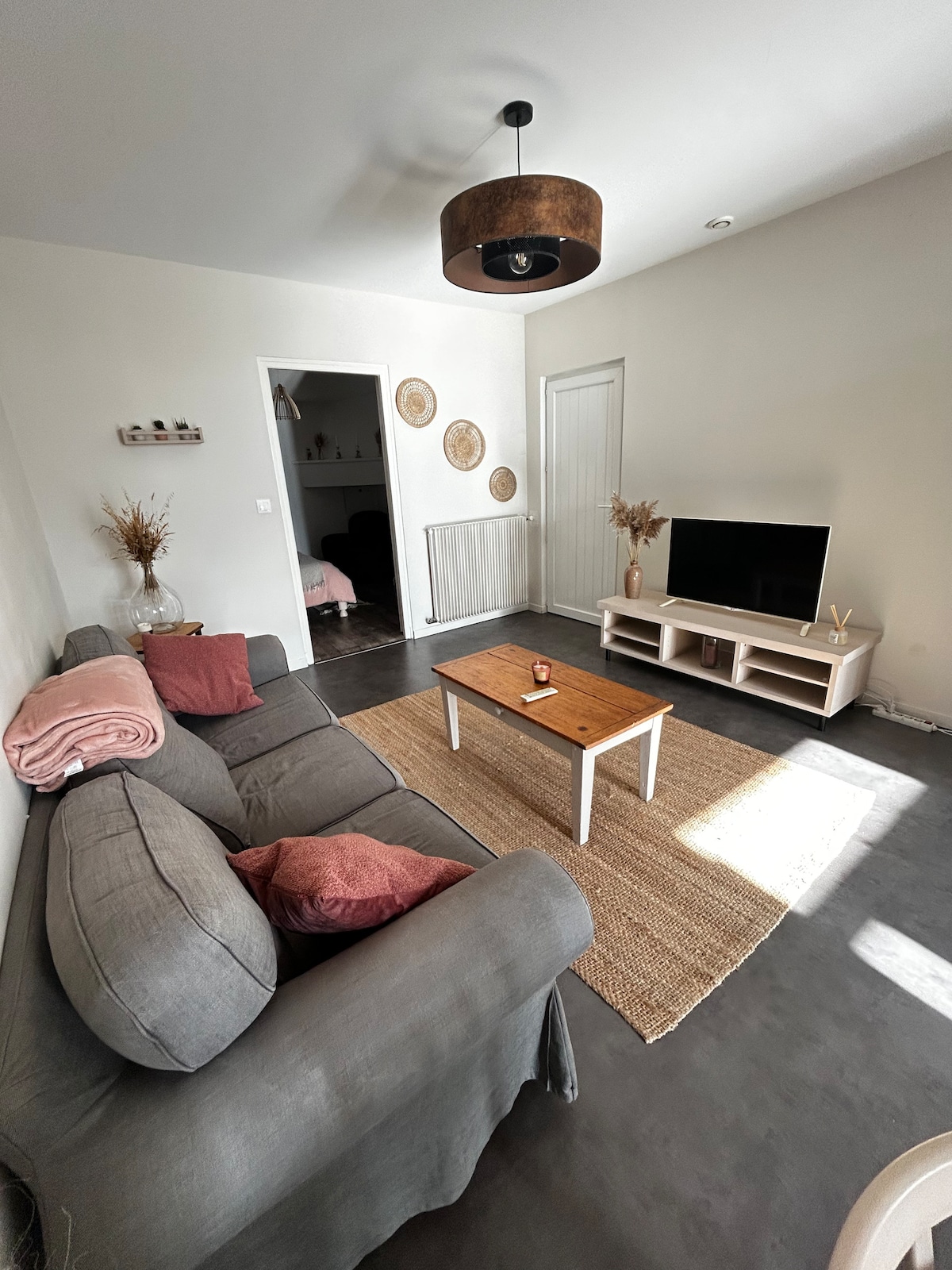 A spacious living area is presented with a comfortable gray sofa adorned with pink cushions. A small wooden coffee table occupies the center, while a flat-screen TV is mounted on a media console. Light enters through an adjacent door, illuminating the neutral-colored walls.