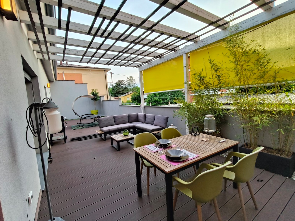 A spacious terrace features a comfortable sofa, dining table, and four chairs. Decorative plants are arranged along the perimeter, and yellow blinds provide shade. A hammock-style chair is positioned in one corner, creating an inviting outdoor space.