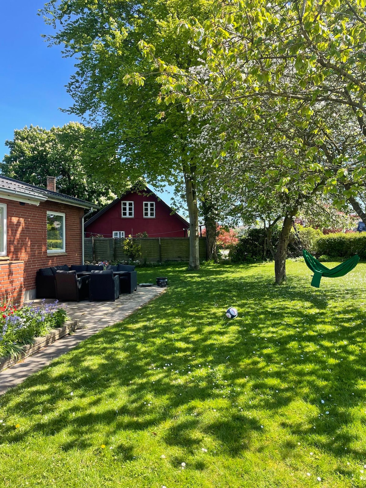 A serene garden area features lush green grass and a variety of trees, including a hammock for relaxation. A patio with seating arrangements is visible, alongside colorful flower beds, while a charming red house can be seen in the background. Bright blue skies enhance the outdoor setting.