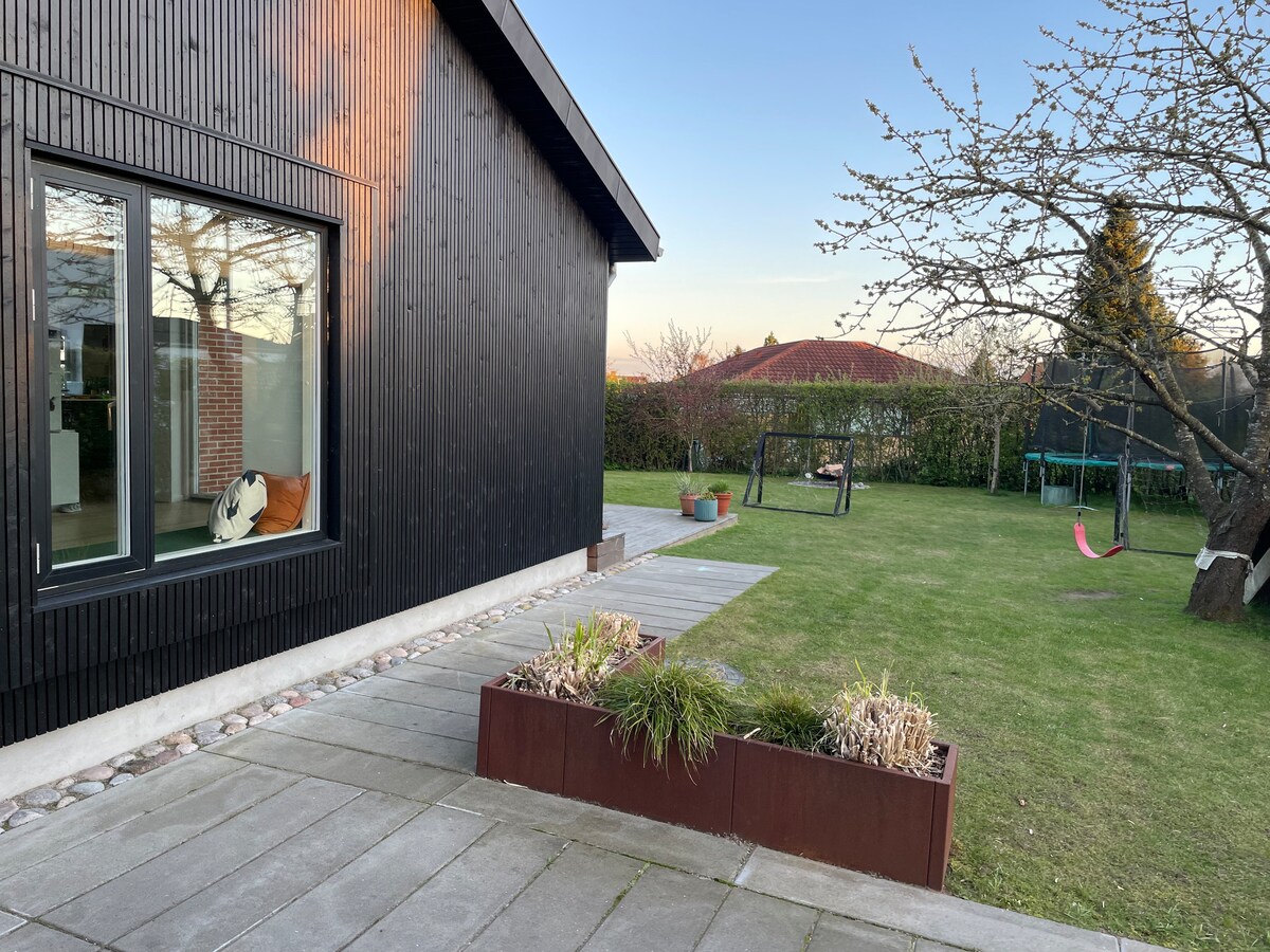 A spacious outdoor area is featured, showcasing a well-maintained lawn surrounded by planted flower beds. A dark wooden house stands in the background, with large windows reflecting the natural light. Play equipment, such as swings and a hammock, is visible, enhancing the family-friendly environment.