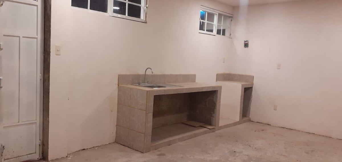 A simple kitchen space is displayed, featuring a built-in countertop with a sink. The walls are painted white, and natural light filters in through multiple small windows above the sink and other areas. The floor appears unfinished, contributing to the minimalist design.