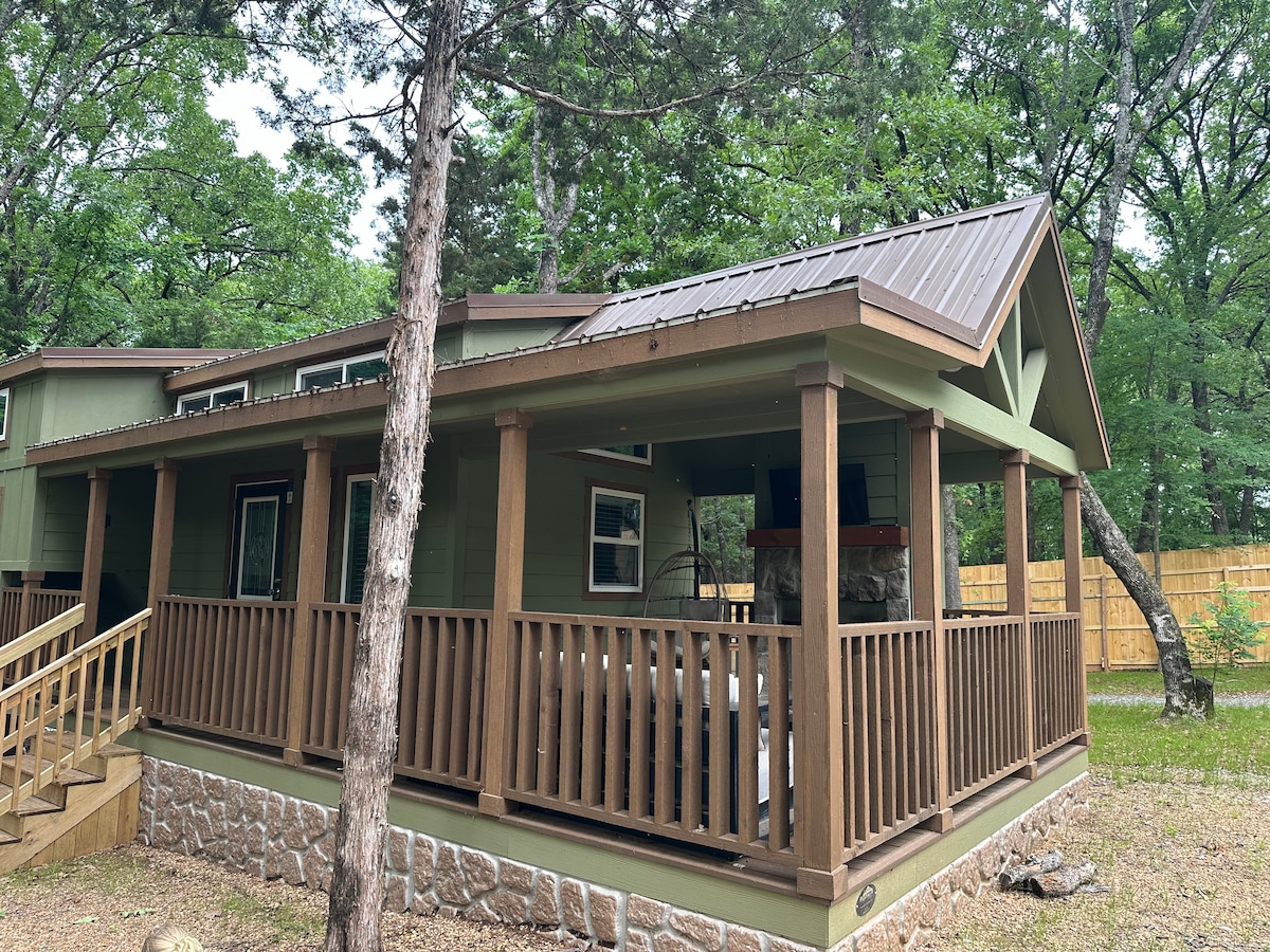 A charming tiny home is set among trees on a gravel path, featuring a covered porch with wooden railings. The sloped roof is complemented by a stone accent wall, and large windows provide natural light to the interior. Surrounding greenery enhances the private setting.