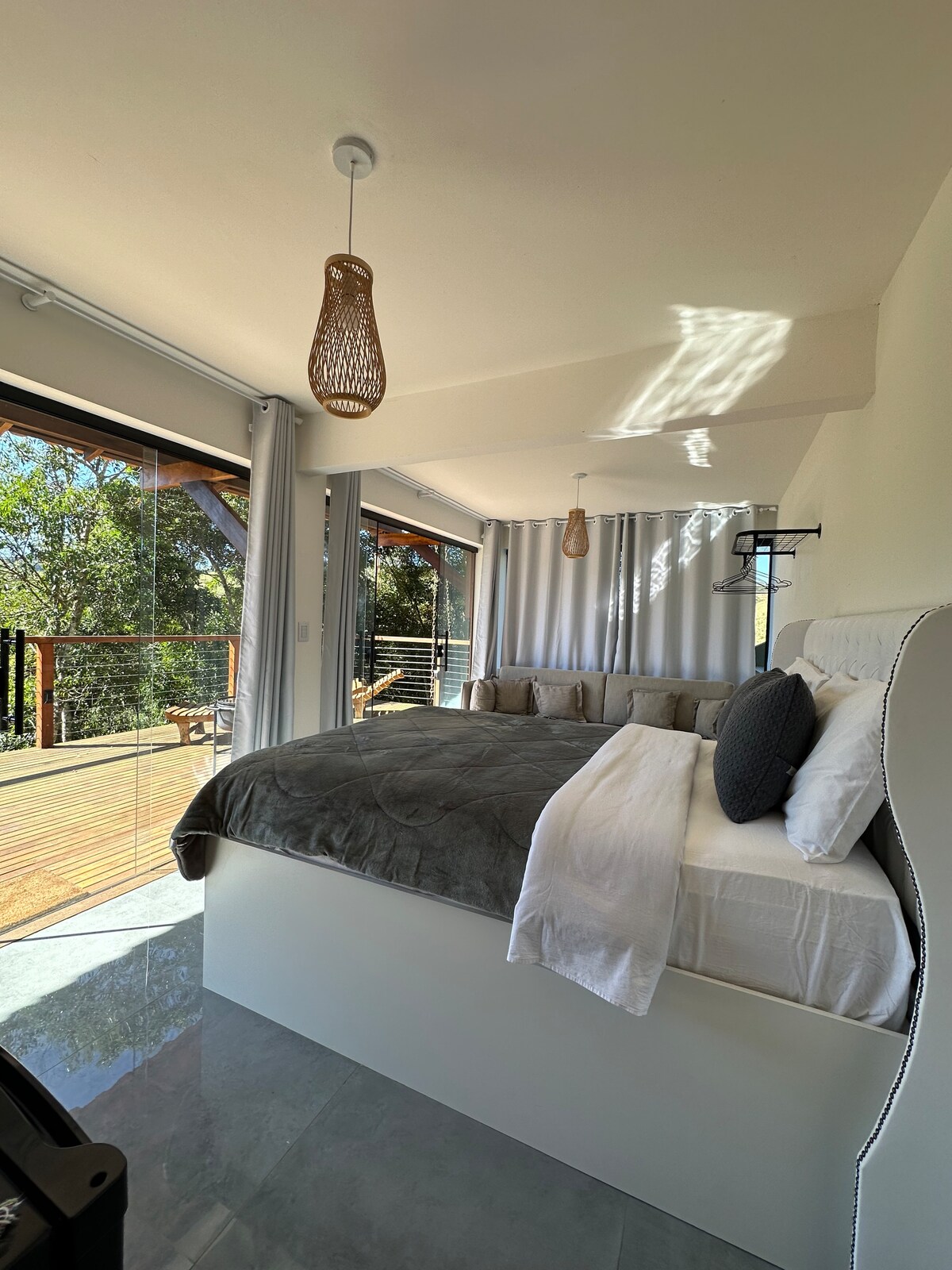 A spacious bedroom features a comfortable bed with neutral bedding and a soft throw. Large glass doors open to a wooden deck surrounded by greenery, allowing natural light to fill the space. A stylish light fixture hangs from the ceiling, enhancing the modern decor.