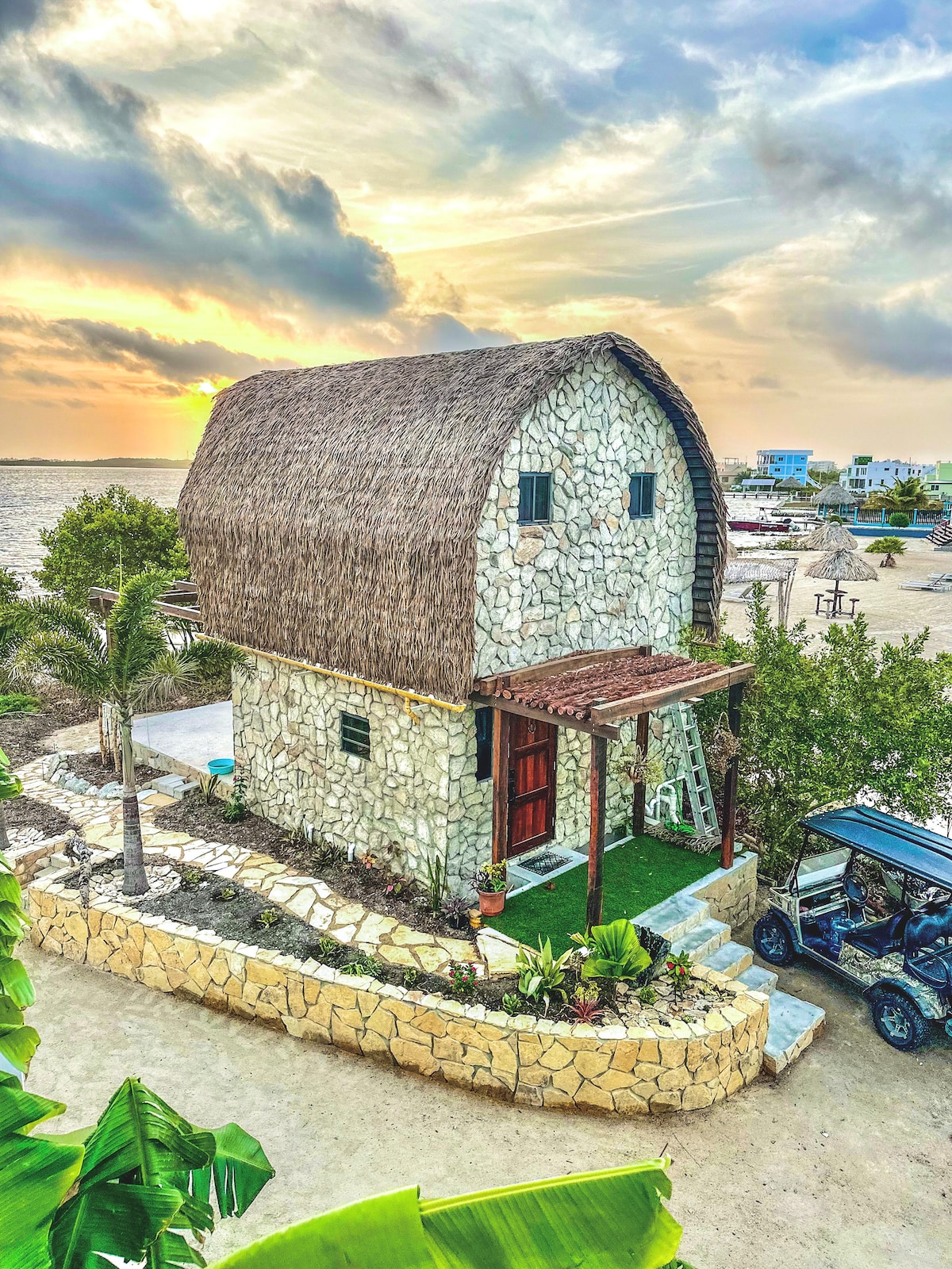 The bungalow’s exterior is constructed from stone and thatch, designed with a distinct curved roof. The entrance is framed by lush plants and a pathway leading to the door, with a golf cart parked nearby. A sunset casts a warm glow over the scene.