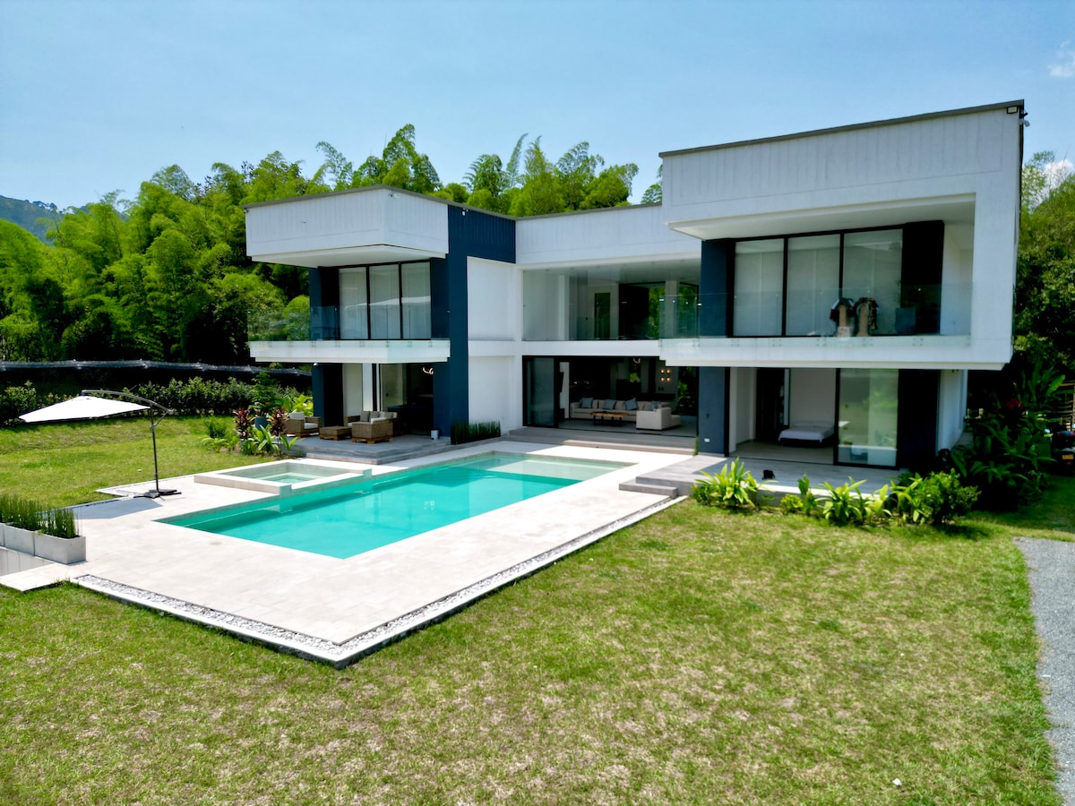 The modern two-story house features large glass doors and a spacious terrace overlooking a swimming pool. Lush greenery surrounds the pool area, while outdoor seating is arranged for relaxation. A shaded umbrella provides comfort, completing the inviting outdoor space.