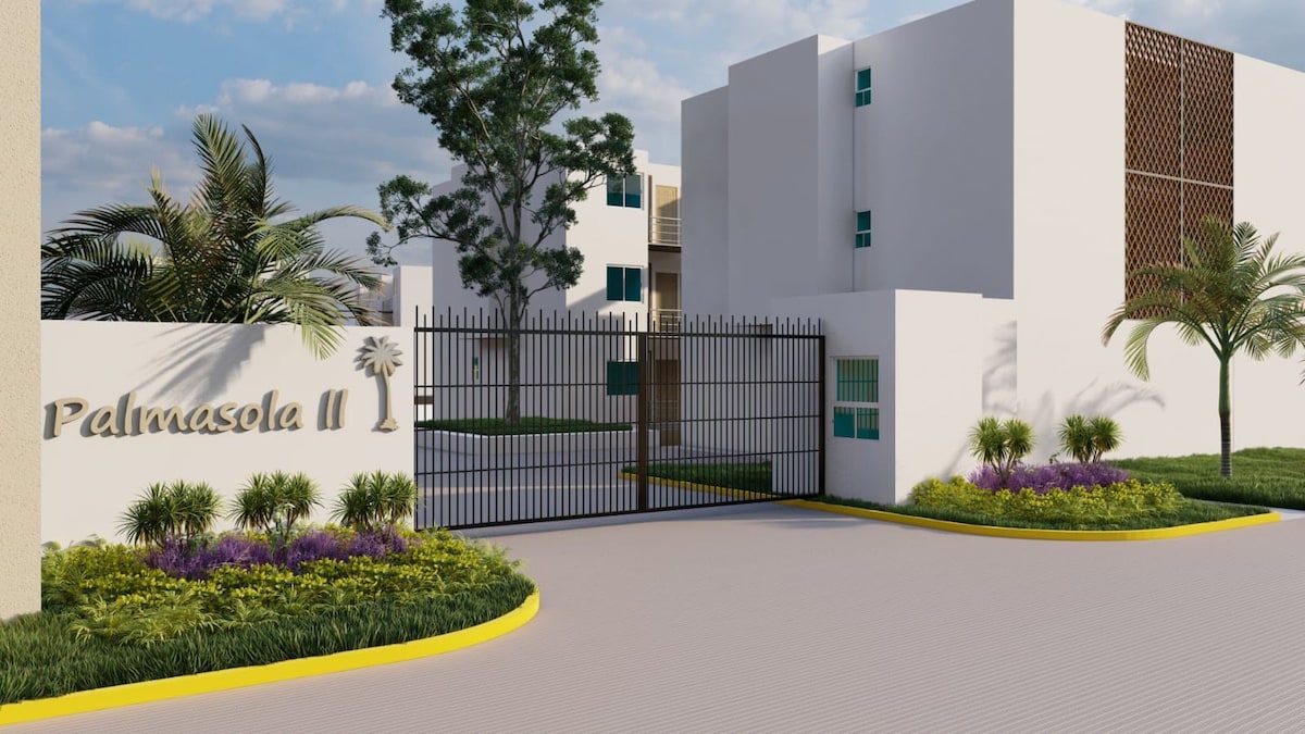 The entrance to Palmasela II is showcased with a secure gate and landscaped grounds. Lush greenery and flowering plants border the pathway, enhancing the welcoming atmosphere. The contemporary architecture of the buildings is visible, featuring a mix of white walls and decorative elements.