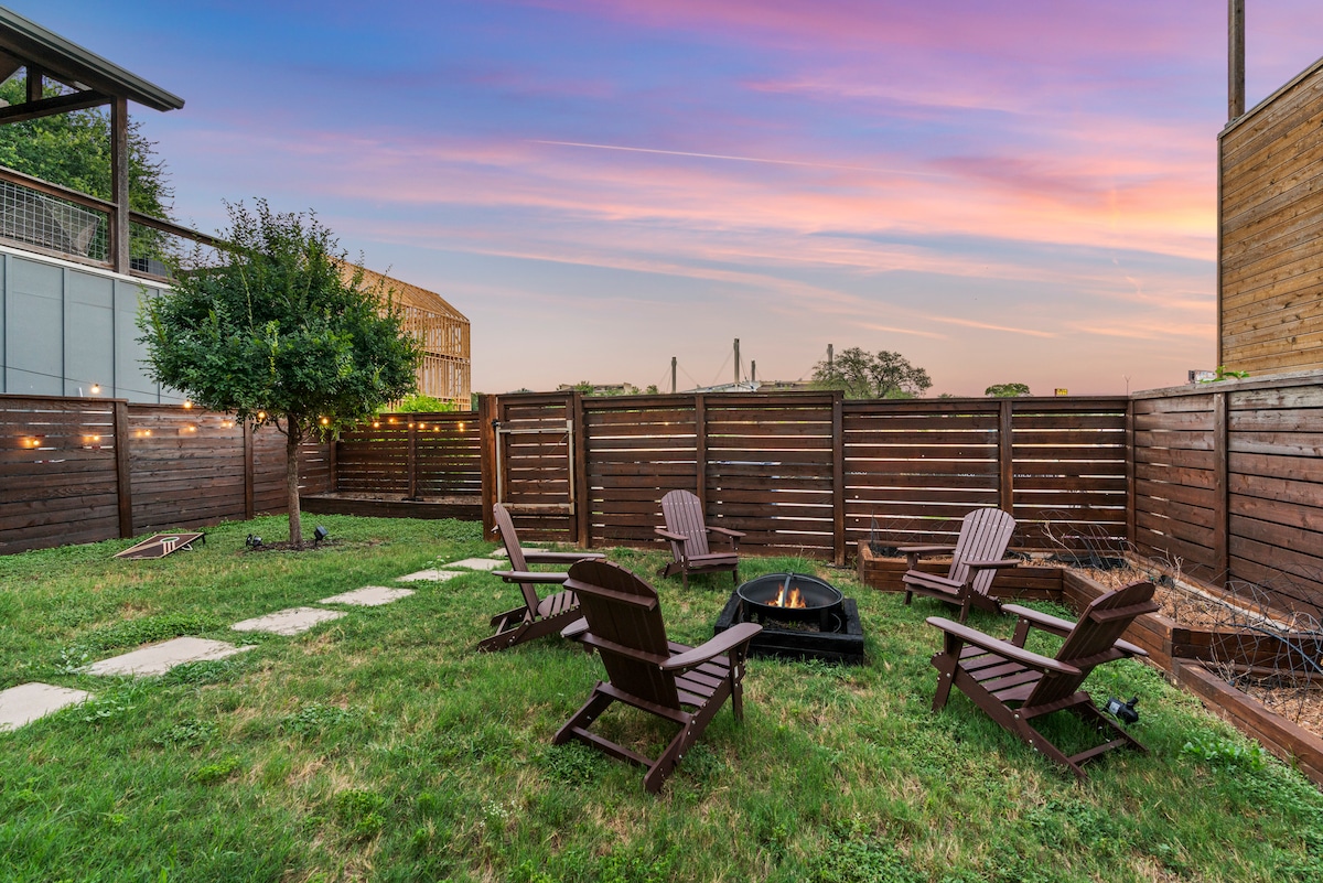 An outdoor space features a cozy firepit surrounded by several camping chairs. Pathways of stepping stones lead through a well-maintained lawn, with soft lighting highlighting a fence that frames the area. A serene sunset sky adds a calming backdrop.