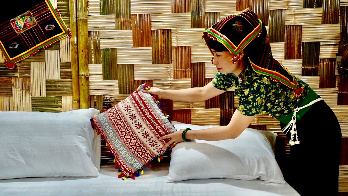 A cozy sleeping area is adorned with decorative pillows featuring vibrant patterns. The walls are crafted from natural materials, creating a warm, earthy atmosphere. A person is seen adjusting a colorful pillow on the neatly made bed, emphasizing a blend of cultural design and comfort.