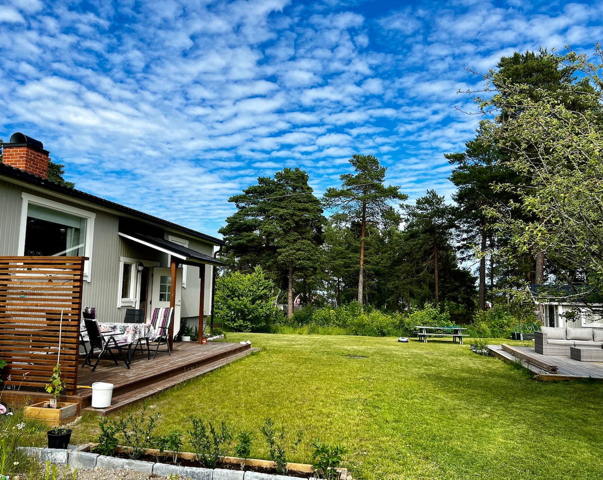 A spacious garden area is shown with well-maintained grass and trees in the background. A patio is visible with a dining table set for outdoor meals, along with a relaxed seating arrangement. The clear blue sky features scattered clouds, creating a peaceful atmosphere.