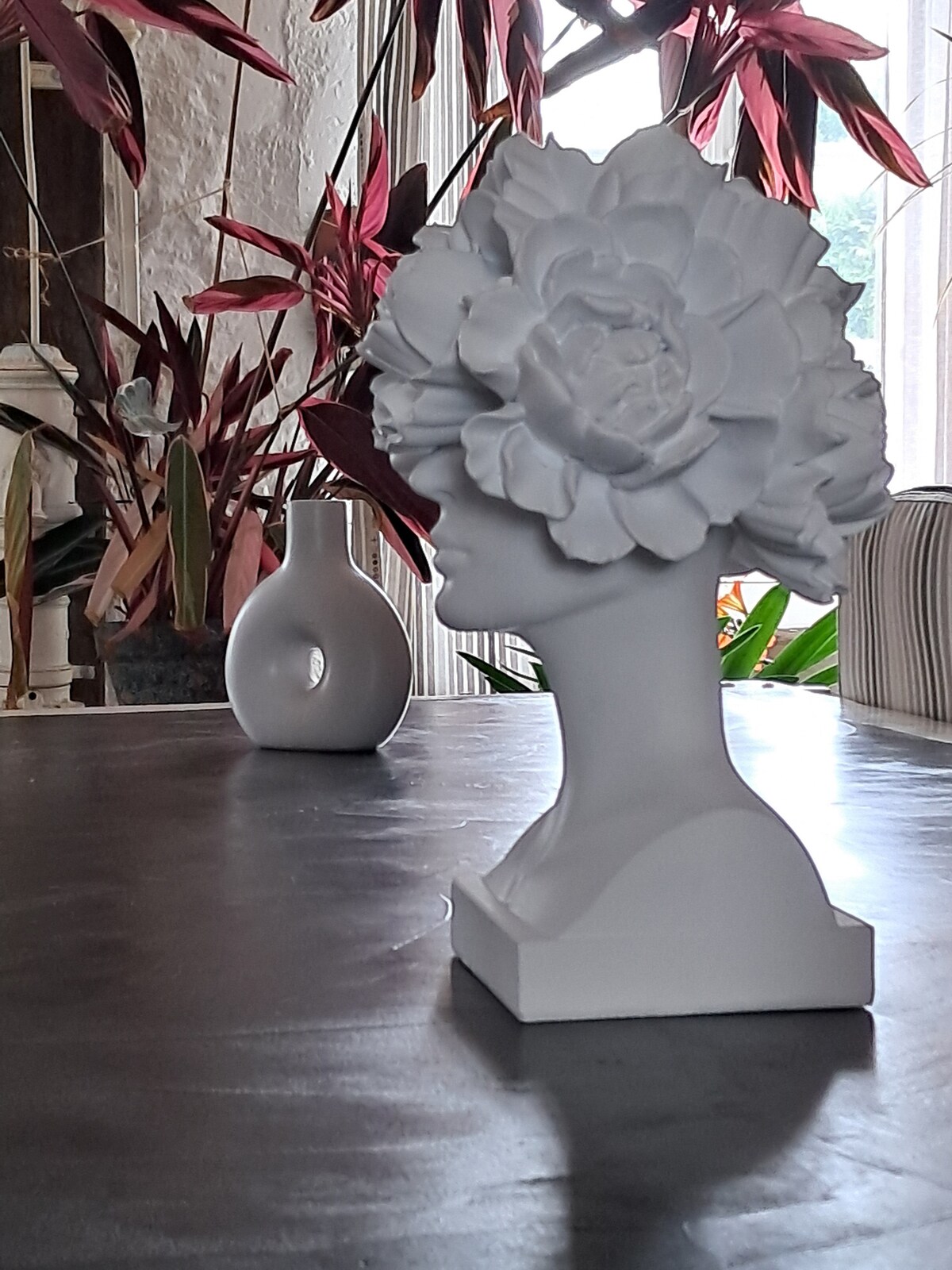 A white bust sculpture with an oversized flower arrangement dominates the foreground. The surrounding area features green foliage and a simple, elegant vase. Soft natural light filters through, enhancing the serene atmosphere created by the minimalist decor.