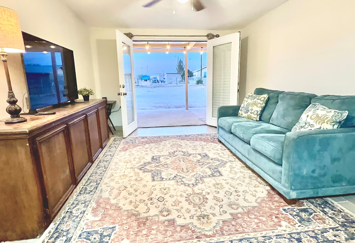 A cozy living area is presented with a soft blue sofa, adorned with decorative pillows. A large area rug with intricate patterns covers the floor. Double doors open to an outdoor space, allowing natural light to fill the room, with a TV and console visible in the background.
