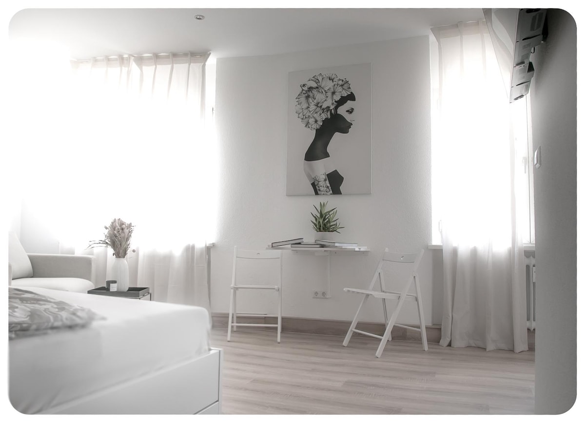 A bright and airy apartment space features large windows with sheer curtains allowing natural light. A minimalist desk is positioned near a wall with art depicting a woman in profile. Two simple white chairs are placed beside the desk, contributing to the uncluttered ambiance.