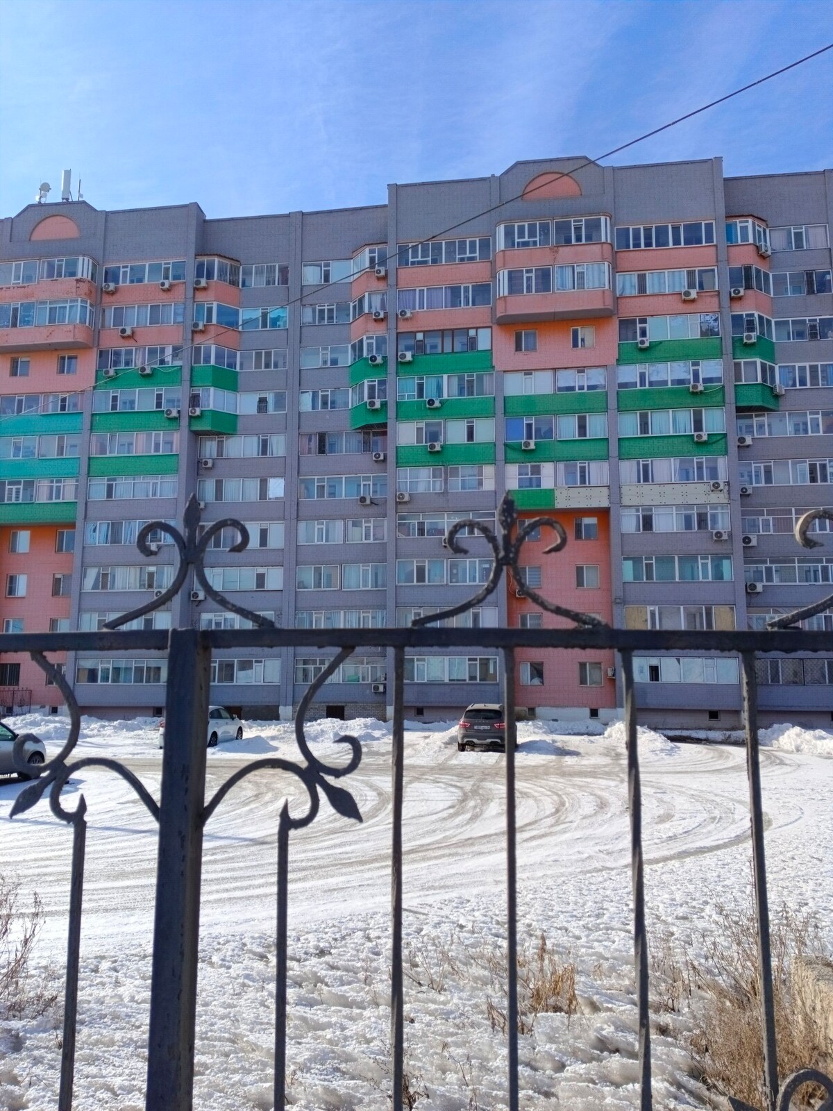 A modern apartment building is visible, characterized by colorful balconies in shades of green and orange. The structure is surrounded by a snow-covered landscape, with a gated entrance in the foreground. Several parked vehicles are visible in the open space.