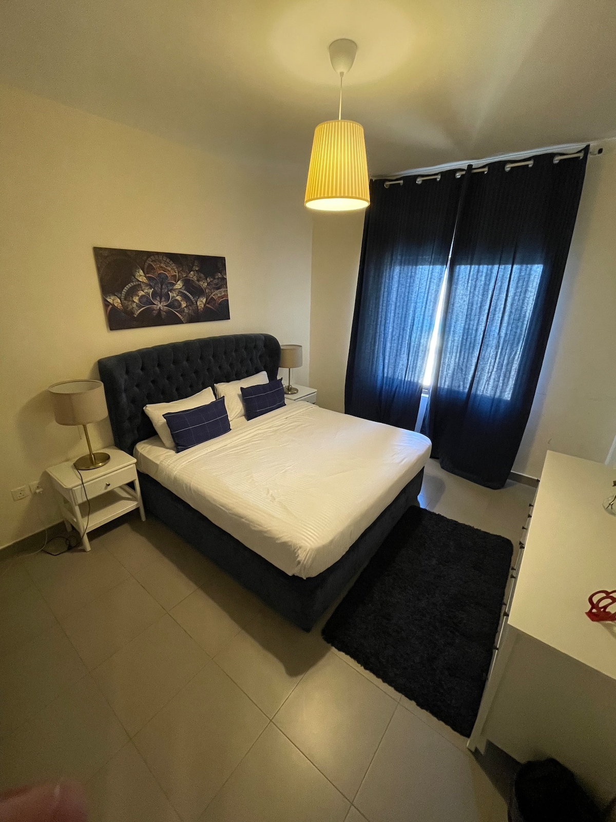 A well-appointed bedroom features a tufted dark headboard with coordinating blue and white pillows placed on a spacious bed. Natural light is allowed in through a large window, draped with dark curtains. A bedside table and a white dresser are present in the room.