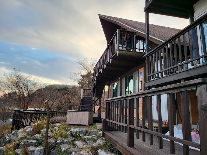 The exterior of a two-story log cabin features a wooden balcony and staircase leading to the upper level. Lush greenery and rocks are situated around the base, while a hot tub is visible on the lower deck. Soft clouds accent the sky in the background.
